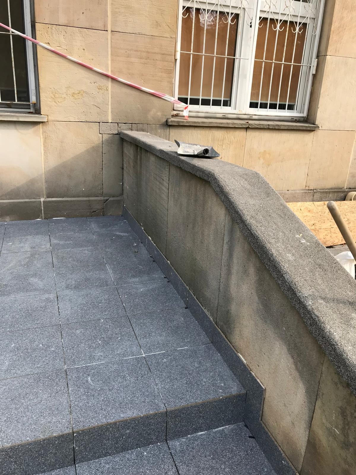 Ułożona szara kostka brukowa na schodach i tarasie przed budynkiem, z widoczną taśmą ostrzegawczą i narzędziem glazurniczym na murku.