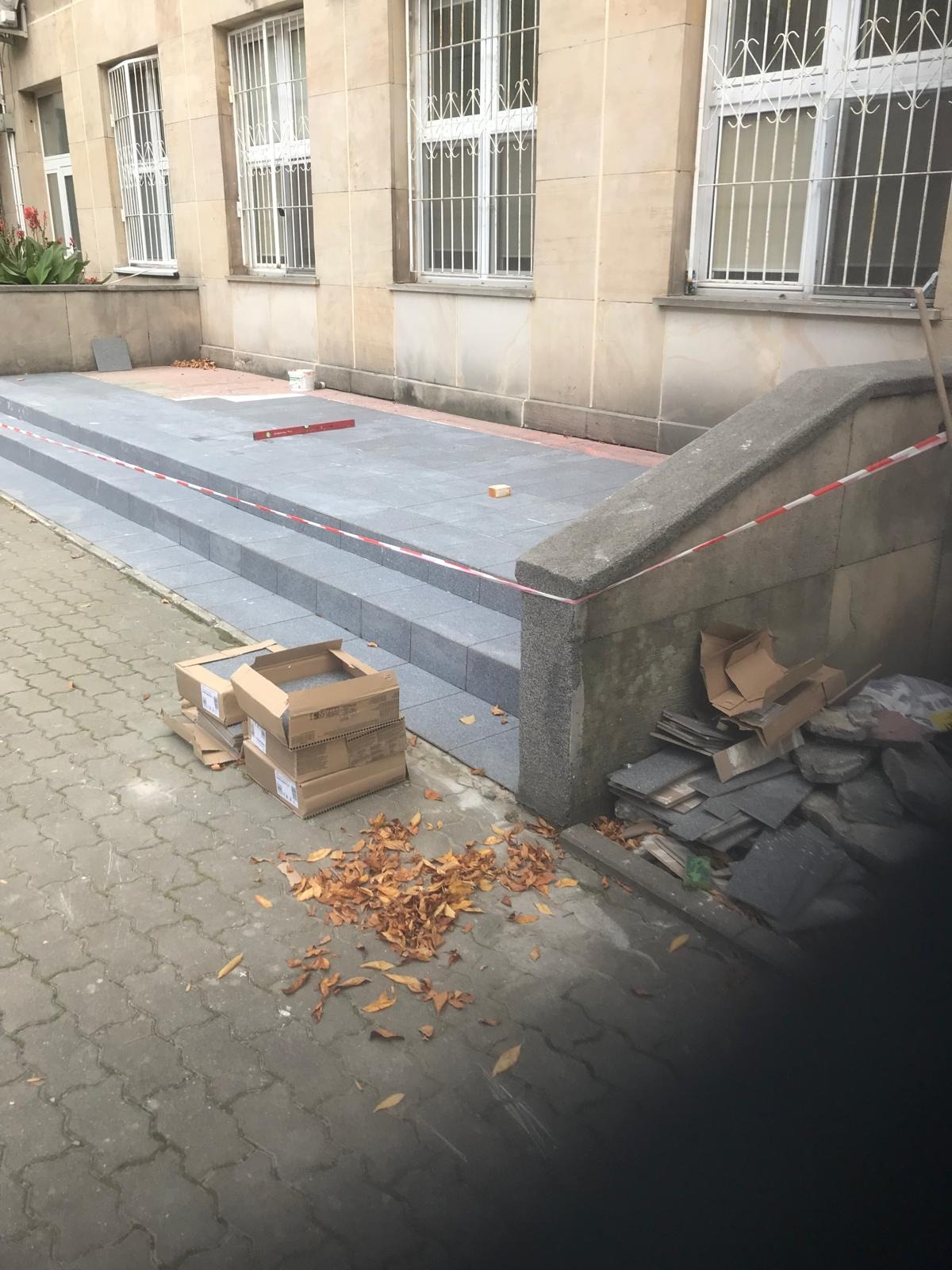 Trwa układanie szarych płyt chodnikowych na schodach wejściowych do budynku, widoczne poziomica, kartony z materiałem i zabezpieczająca taśma ostrzegawcza.