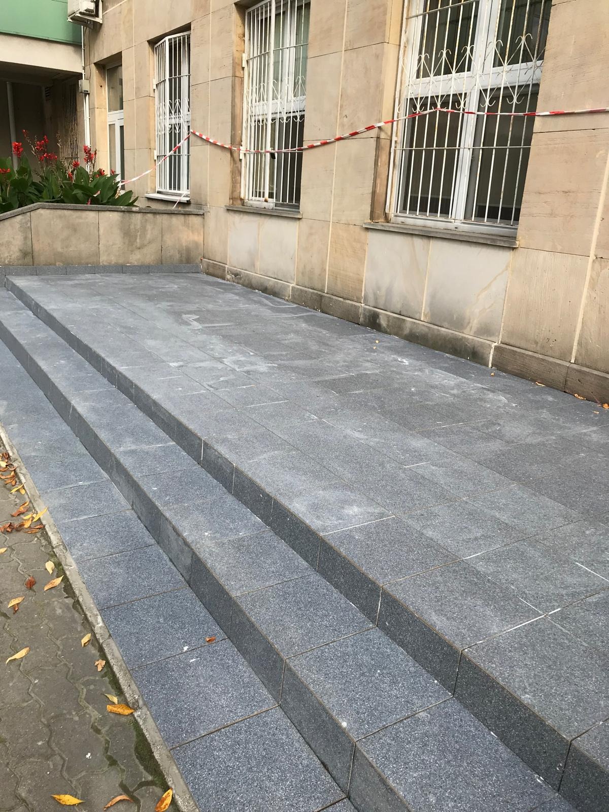 Schody wejściowe wyłożone szarą, granitową kostką, prowadzące do budynku z beżową elewacją i oknami z kratami, zabezpieczone taśmą ostrzegawczą.