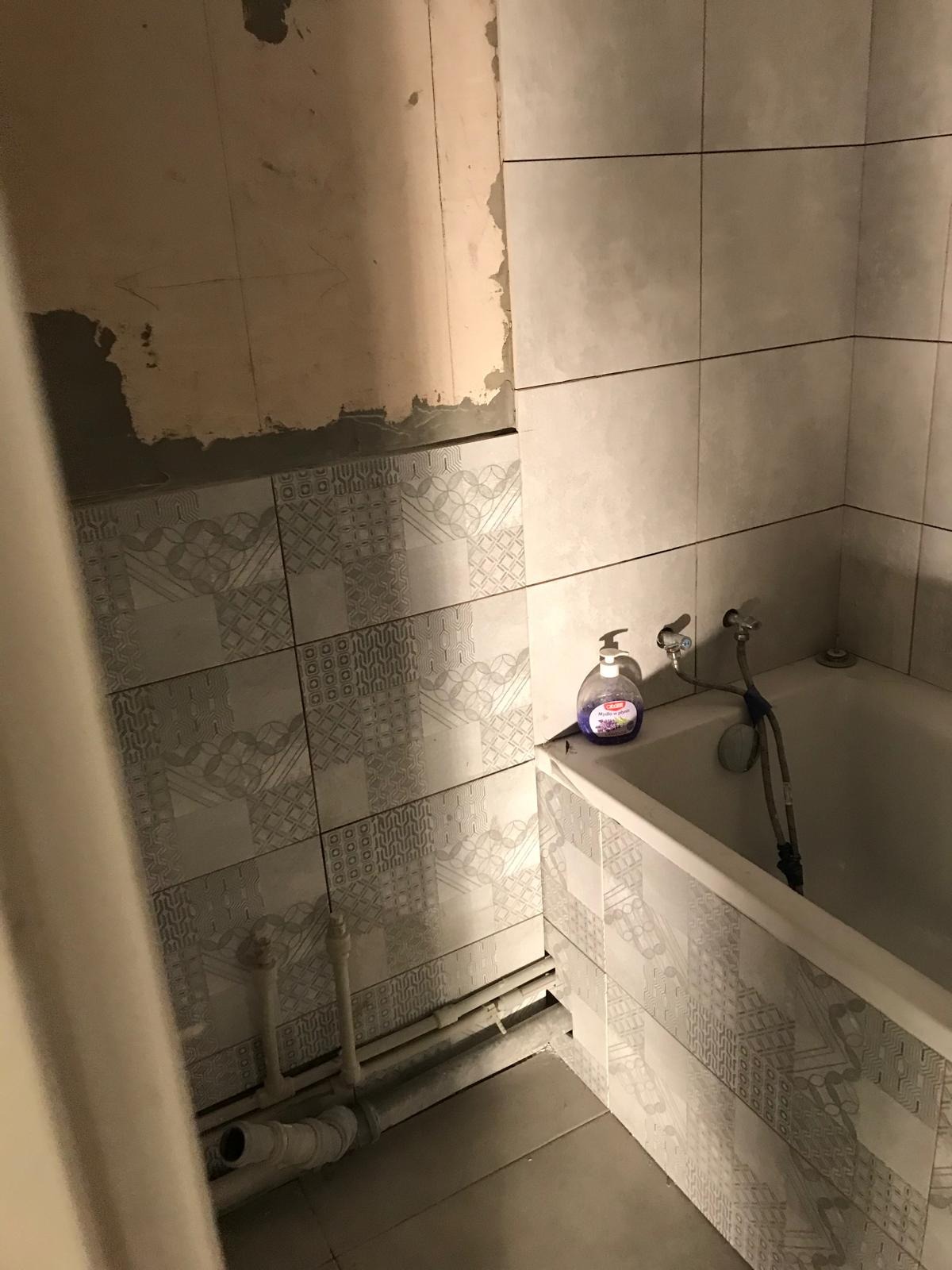 Wnętrze łazienki w trakcie remontu; widoczne częściowo położone płytki z wzorem, fragment ściany przed wykończeniem, rury instalacyjne i butelka mydła w płynie na wannie.