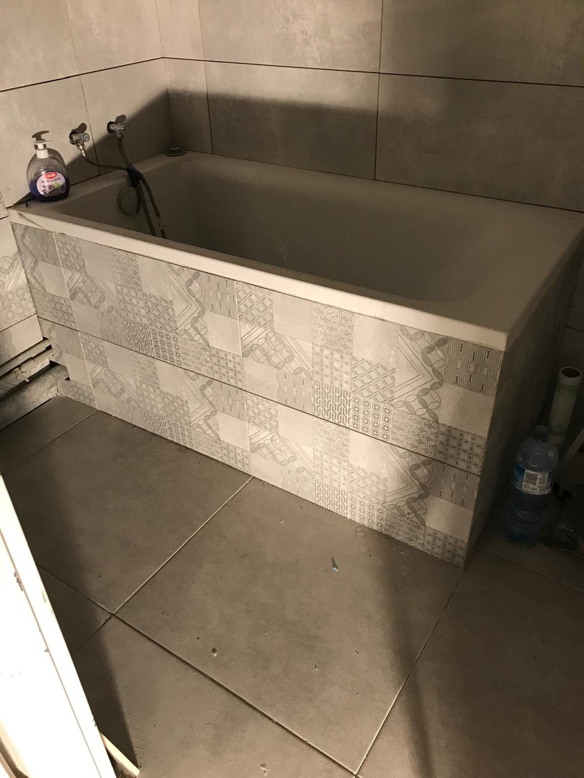 Biała wanna z obudową wyłożoną płytkami w geometryczne wzory, widoczne szare płytki na ścianach i podłodze, butelka płynu do mycia na rancie wanny, niedokończone wykończenie przy obudowie wanny.