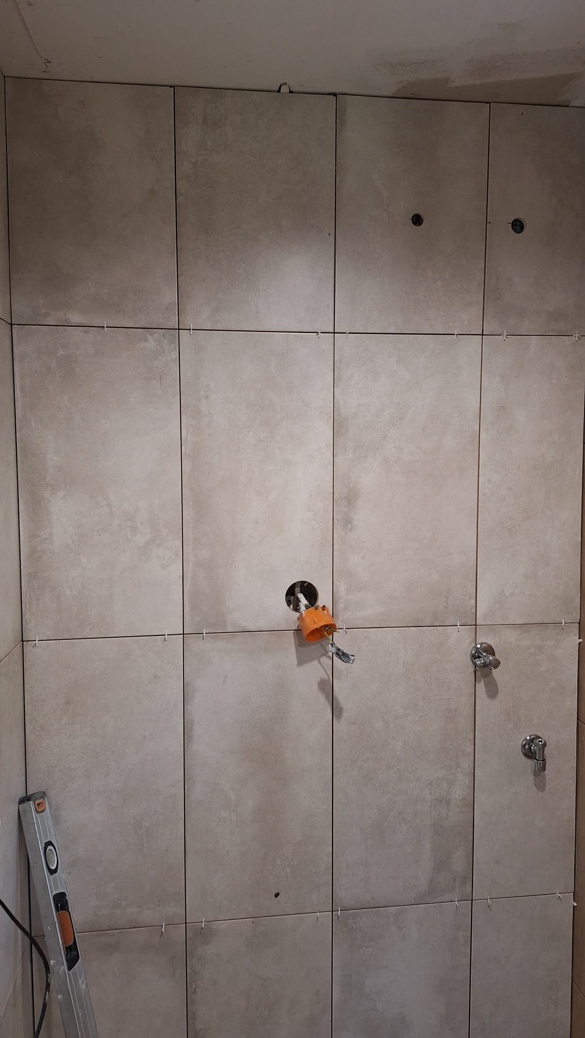 Ściana w trakcie wykańczania płytkami ceramicznymi w łazience, widoczne krzyżyki dystansowe, poziomica przy ścianie, puszka elektryczna i armatura.