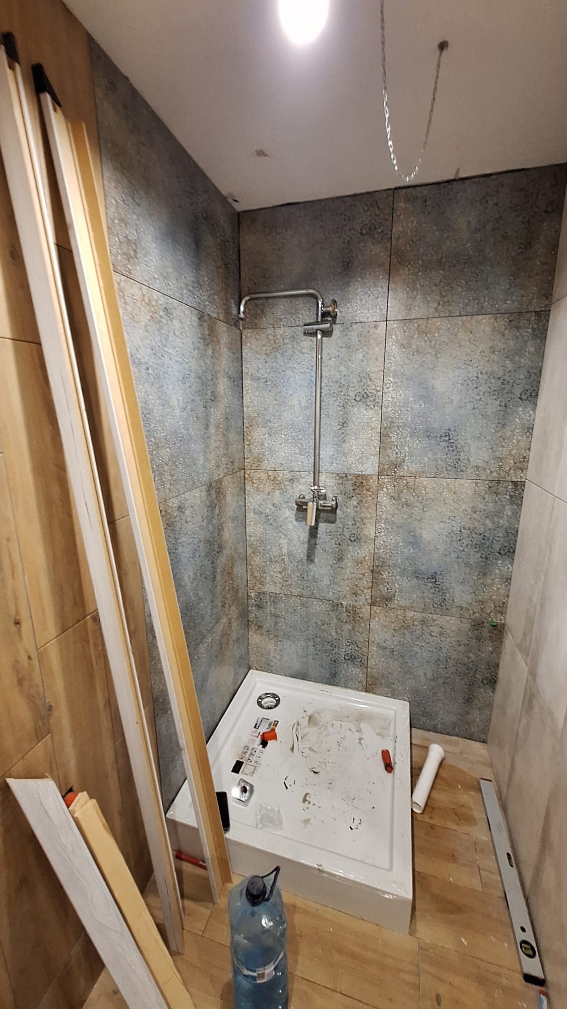 Wnętrze łazienki w trakcie remontu: zamontowany brodzik, surowa instalacja prysznicowa, płytki w odcieniach szarości i brązu na ścianach, listwy przypodłogowe oparte o ścianę, butelka z wodą...