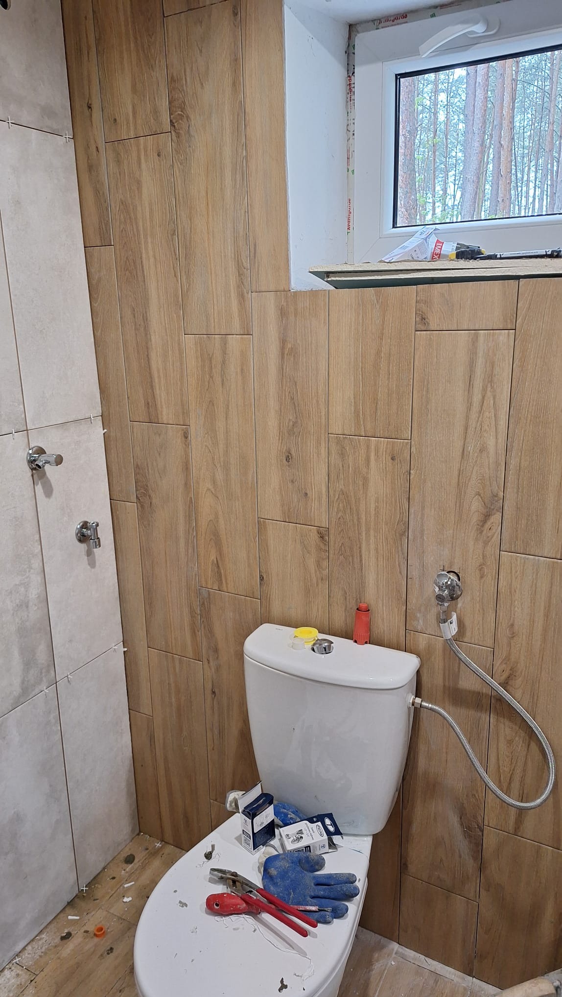 Łazienka w trakcie wykańczania: częściowo ułożone płytki imitujące drewno na ścianach, biała toaleta z niebieskimi rękawicami roboczymi i narzędziami na desce, widok przez okno na las.