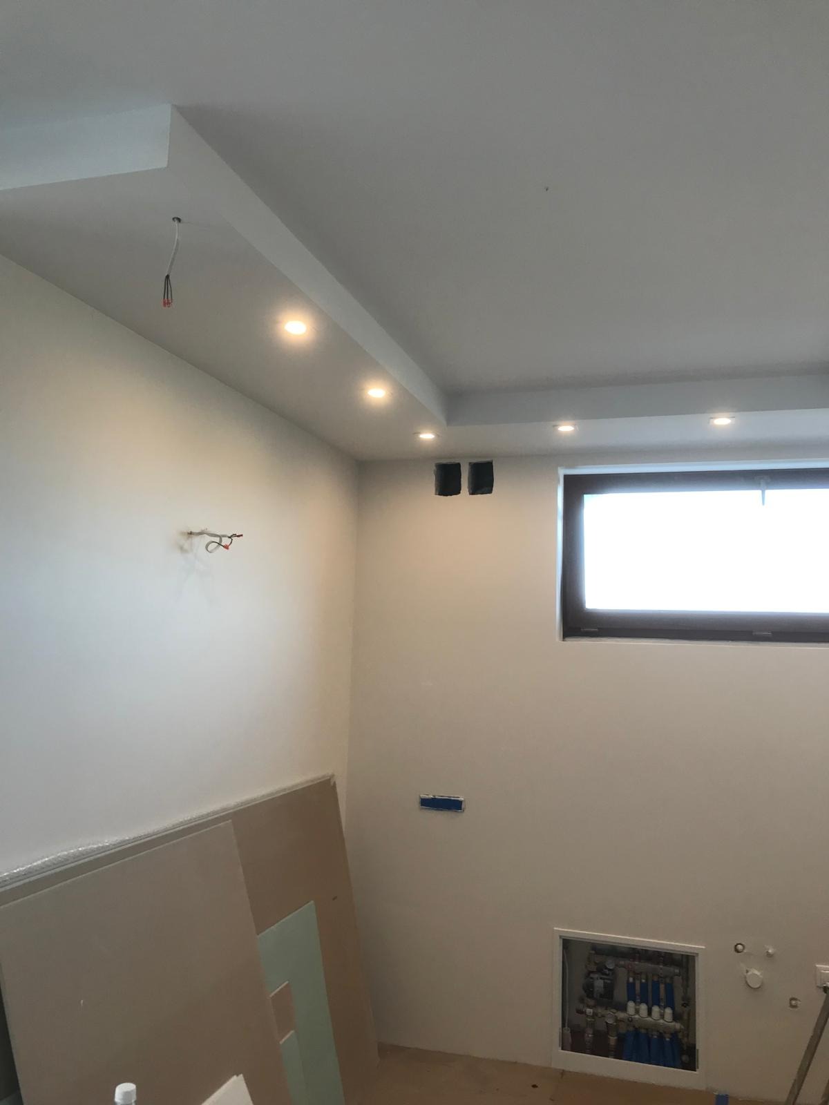 Wnętrze w trakcie prac wykończeniowych: białe ściany po malowaniu, podwieszany sufit z oświetleniem punktowym, widoczne przewody, okno z ciemną ramą, płyty gipsowo-kartonowe oparte o ścianę...