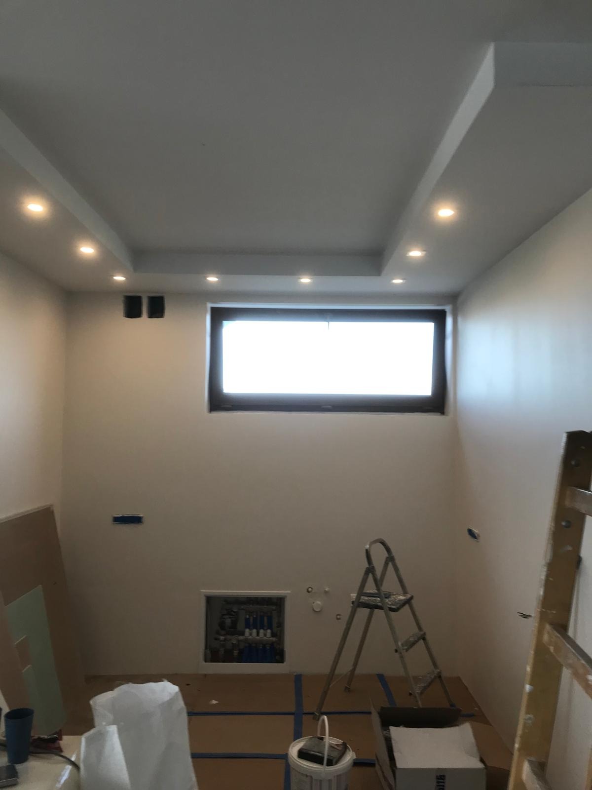 Wnętrze w trakcie prac wykończeniowych: świeżo pomalowane ściany na biało, widoczne maskownice głośników w suficie podwieszanym z oświetleniem punktowym, okno z ciemną ramą, zabezpieczona podłoga...