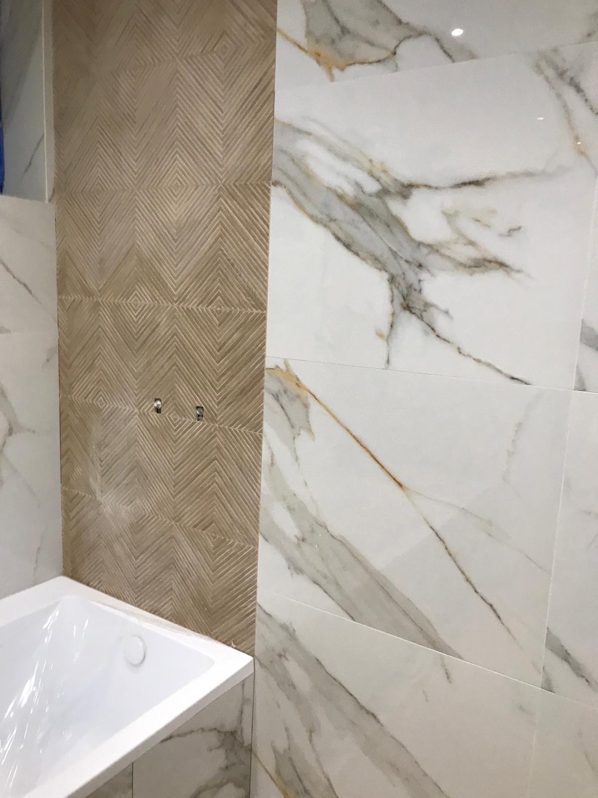Wykończona łazienka z wanną, ściany wyłożone płytkami imitującymi marmur oraz dekoracyjnymi płytkami w kolorze beżowym o wzorze geometrycznym, widoczne dwa metalowe punkty mocowania.