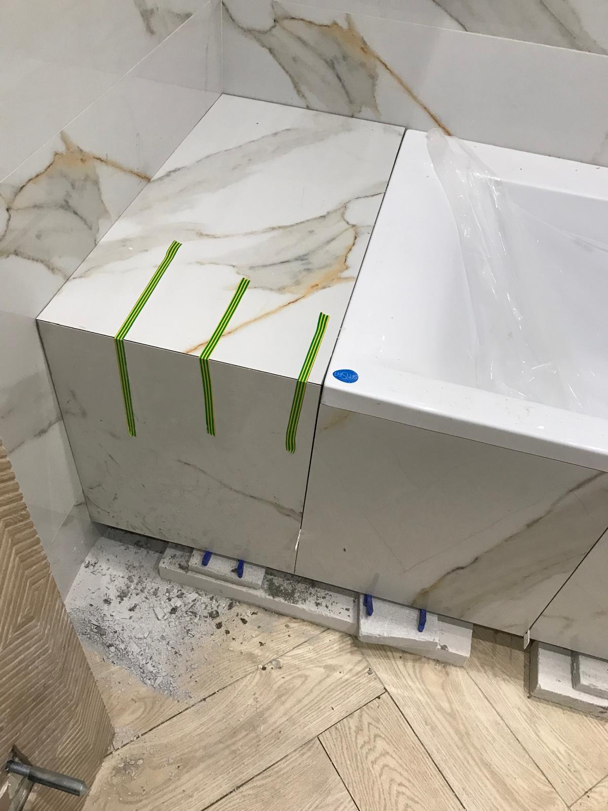 Montaż płytek imitujących marmur na obudowie wanny, z użyciem zielonej taśmy maskującej i poziomujących klinów, widoczny fragment drewnianej podłogi i okładziny ściany.