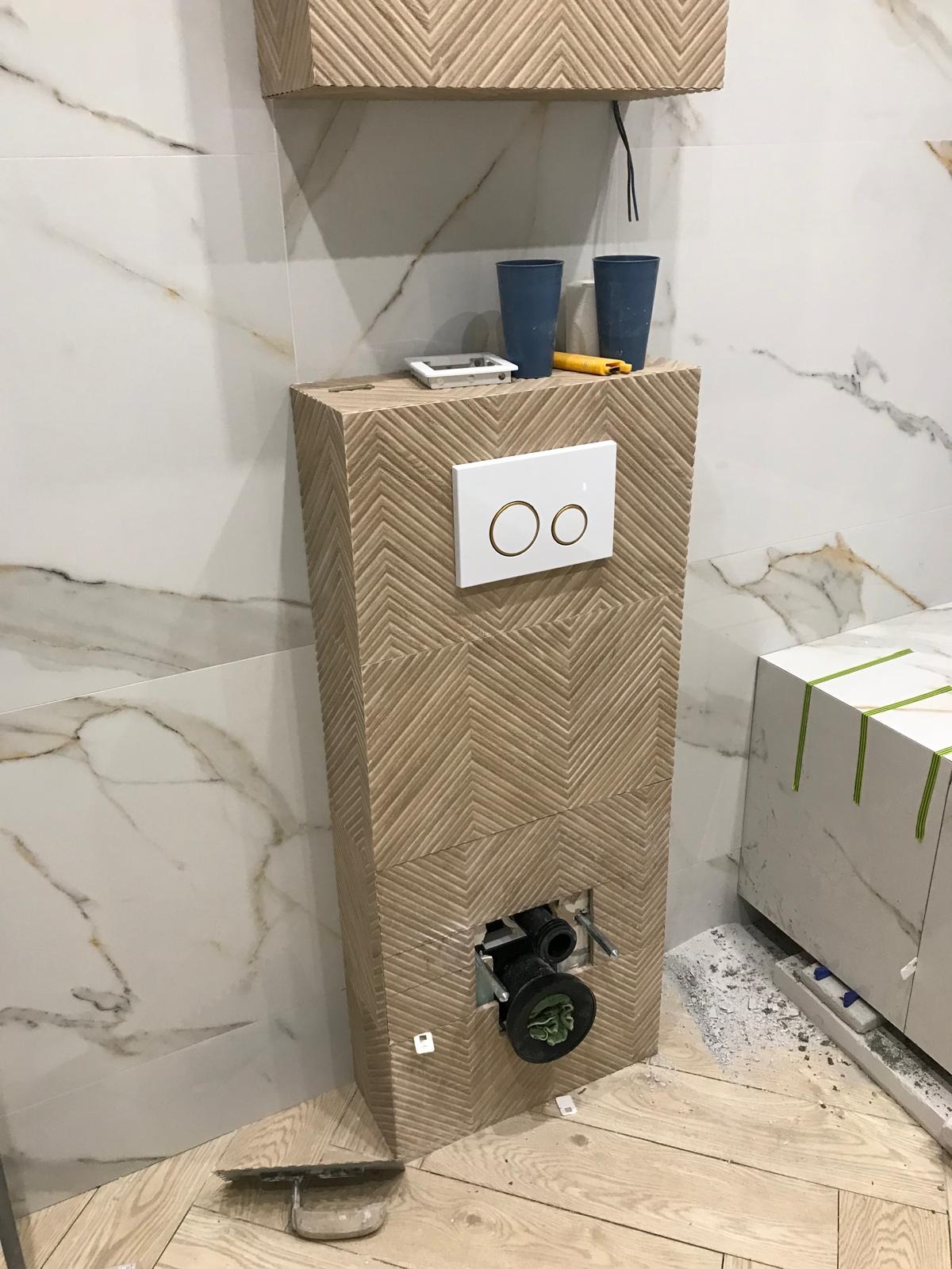 Wiszący stelaż WC obudowany płytkami z wzorem chevron, widoczne przyłącza kanalizacyjne, biała ściana z marmurowym wzorem, podłoga z paneli, niebieskie kubki, nóż i poziomica na górze stelaża.