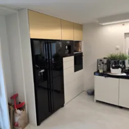 Nowoczesna kuchnia na wymiar z czarną lodówką i złotymi szafkami w Cieszynie. Minimalistyczny design, jasne wnętrze, widoczne sprzęty AGD.