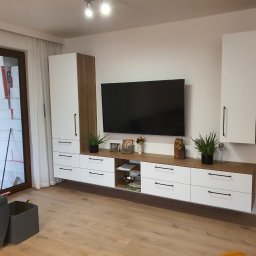 MEBLEZPACZKI.PL PROSTA SPÓŁKA AKCYJNA - Biało-drewniana meblościanka z szufladami i szafkami wiszącymi, z czarnymi uchwytami, pod telewizorem, z dekoracjami i roślinami. Widok na okno z pakietami styropianu.