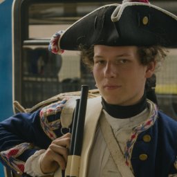 Jakub Bak - Młody mężczyzna w historycznym kostiumie z XVIII wieku, trzymający pałeczki do bębna, na tle autobusu.