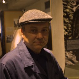 Jakub Bak - Portret mężczyzny w brązowej czapce kaszkietówce i granatowej kurtce na tle wystawy fotograficznej.