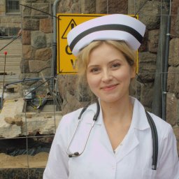Jakub Bak - Uśmiechnięta młoda kobieta w stroju pielęgniarki, z stetoskopem na szyi, pozuje na tle kamiennej ściany i żółtego znaku ostrzegawczego na placu budowy.