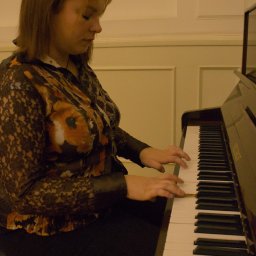 Jakub Bak - Kobieta w kwiecistej bluzce gra na pianinie Petrof w pomieszczeniu z panelami ściennymi.