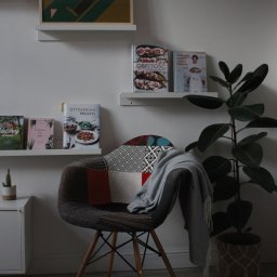 Jakub Bak - Przytulny kącik do czytania z krzesłem patchworkowym, pledem, półkami z książkami kucharskimi i plakatem 'Tu się czyta', dopełniony rośliną doniczkową w plecionym koszu.