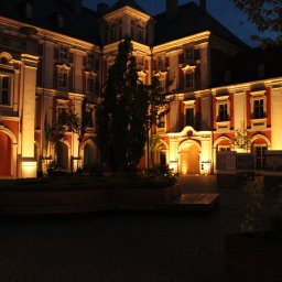 Jakub Bak - Fasada budynku w stylu barokowym, oświetlona ciepłym światłem nocą, z zielenią i brukowaną nawierzchnią na pierwszym planie.
