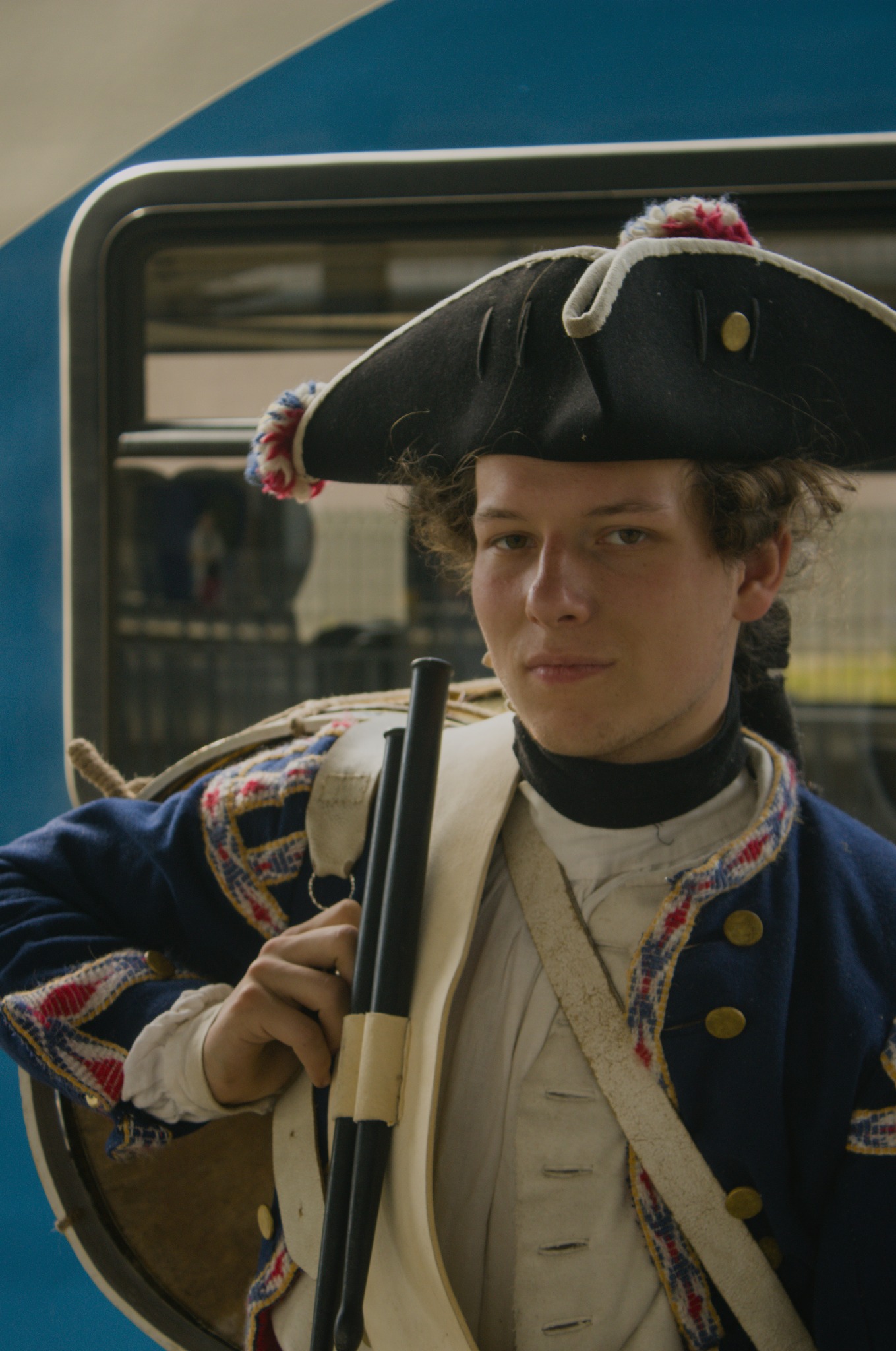 Młody mężczyzna w historycznym kostiumie z XVIII wieku, trzymający pałeczki do bębna, na tle autobusu.