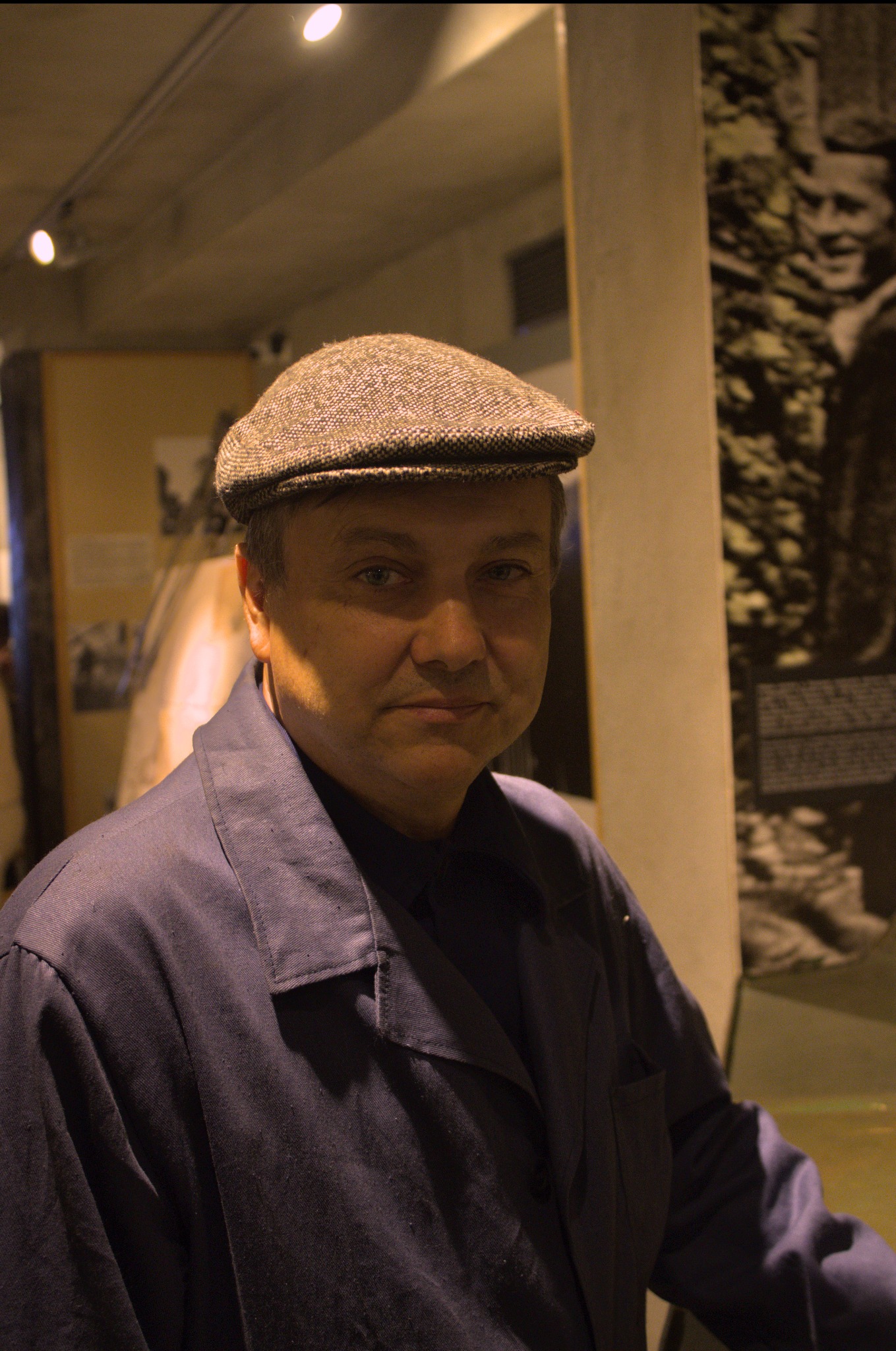 Portret mężczyzny w brązowej czapce kaszkietówce i granatowej kurtce na tle wystawy fotograficznej.