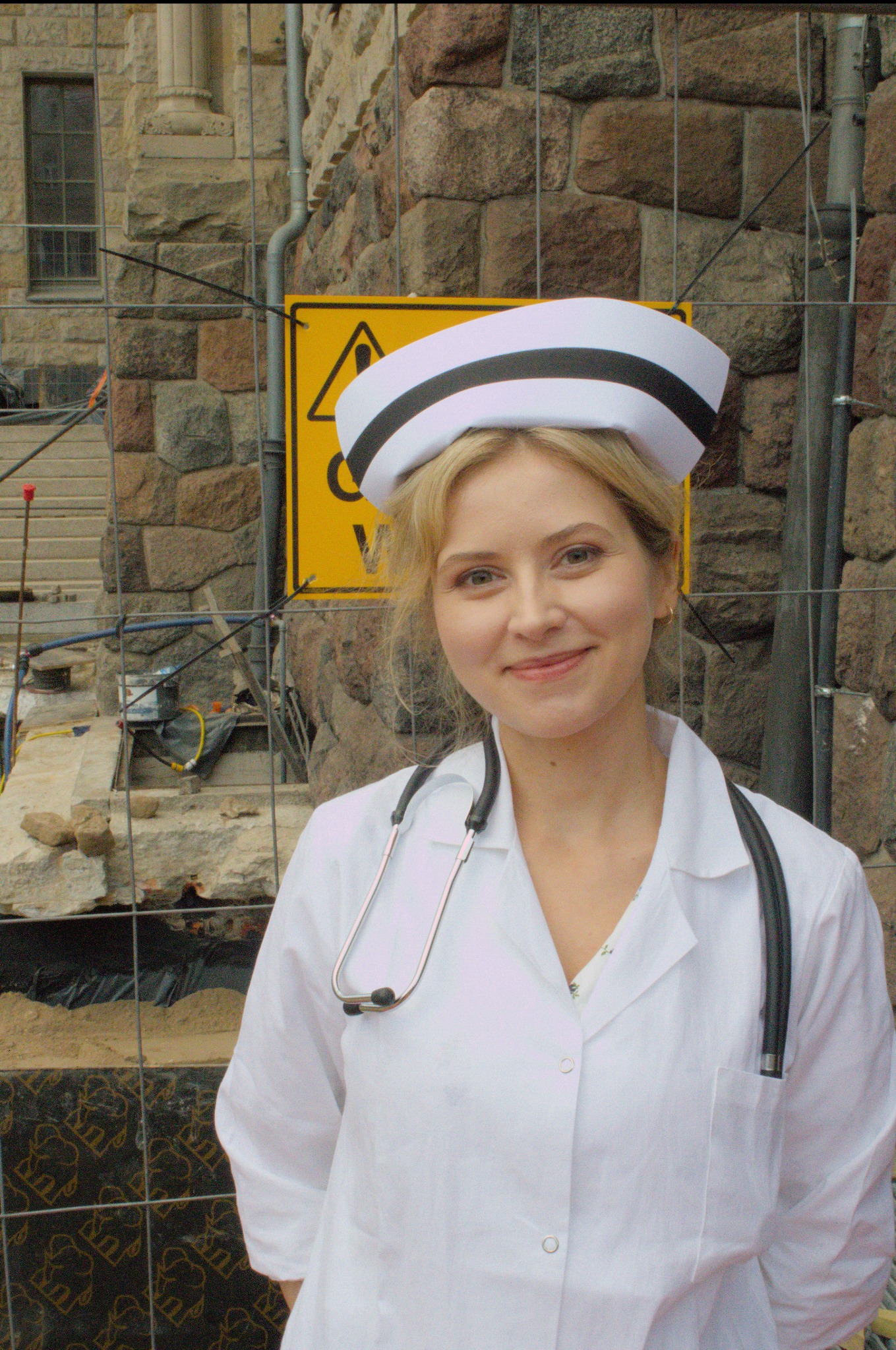 Uśmiechnięta młoda kobieta w stroju pielęgniarki, z stetoskopem na szyi, pozuje na tle kamiennej ściany i żółtego znaku ostrzegawczego na placu budowy.