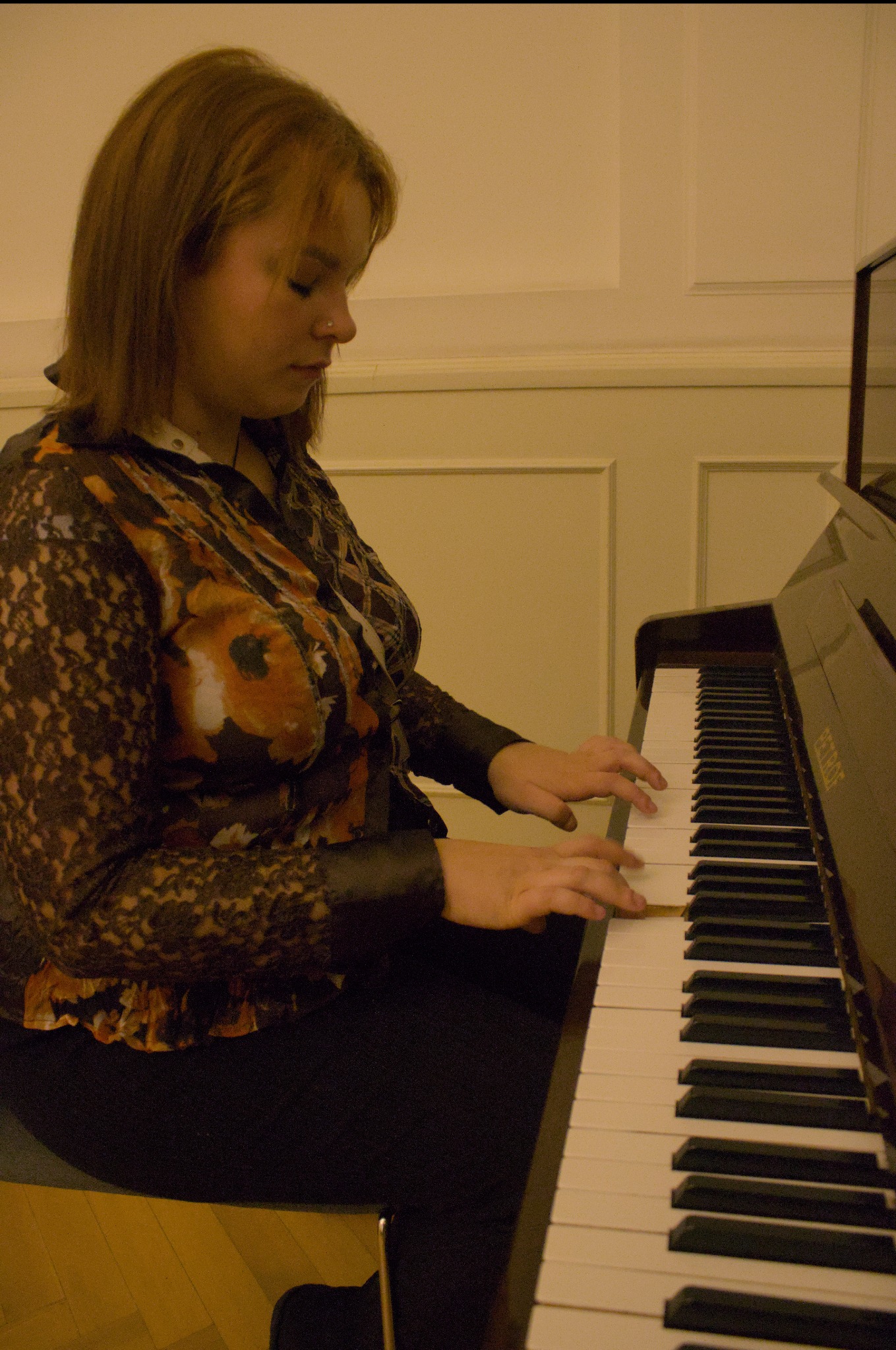 Kobieta w kwiecistej bluzce gra na pianinie Petrof w pomieszczeniu z panelami ściennymi.