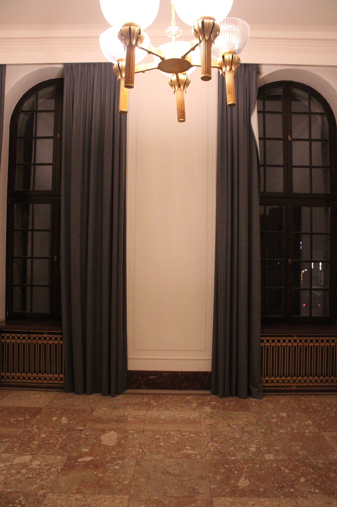 Wnętrze z marmurową podłogą, dwoma łukowatymi oknami z ciemnymi ramami zasłoniętymi szarymi zasłonami i dekoracyjnym, złotym grzejnikiem pod każdym oknem, oraz modernistycznym żyrandolem z białymi...
