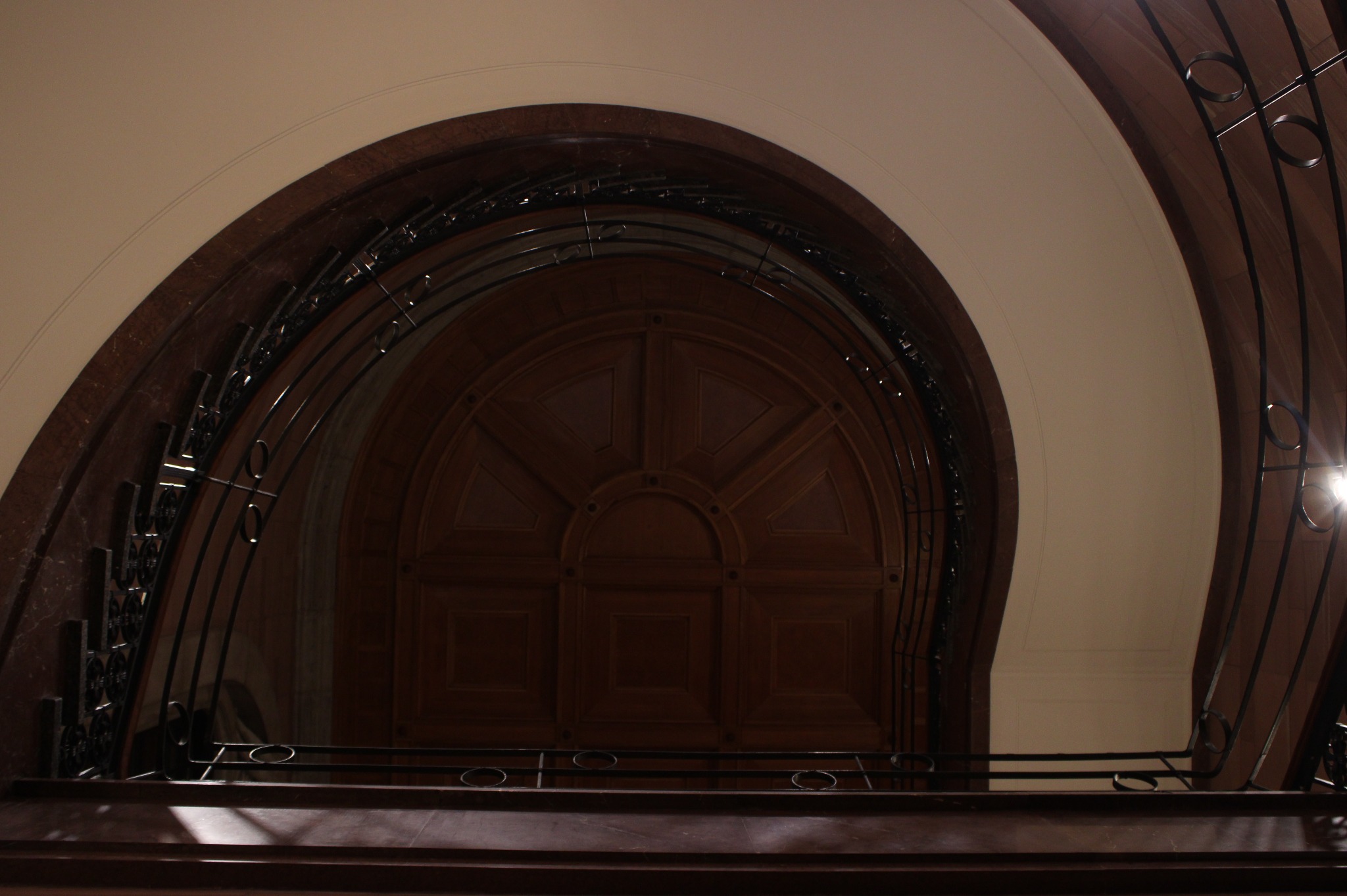 Wnętrze klatki schodowej z ciemnego drewna, z widoczną półokrągłą, zdobioną ścianą na końcu biegu schodów i metalową balustradą z okrągłymi elementami.