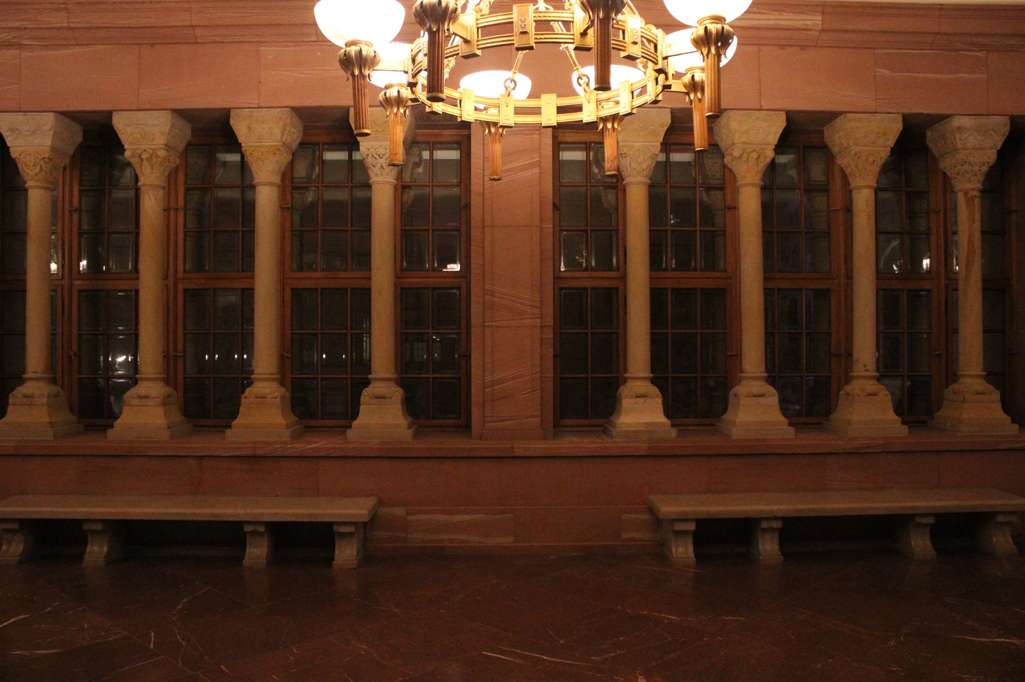 Wnętrze holu z marmurową podłogą, kolumnami, oknami z kratami i ozdobnym żyrandolem.