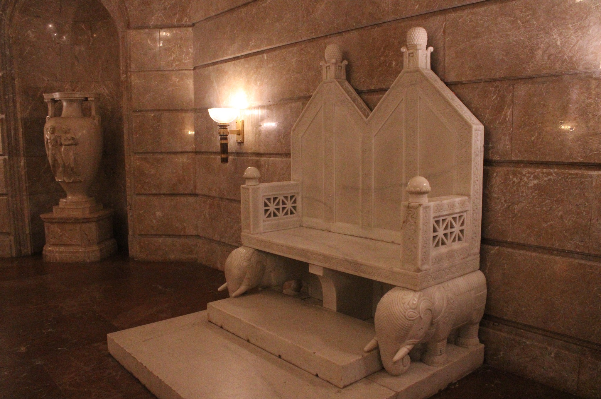 Monumentalna marmurowa ławka z motywem słoni w klasycznym wnętrzu z marmurowymi ścianami i wazą w tle.