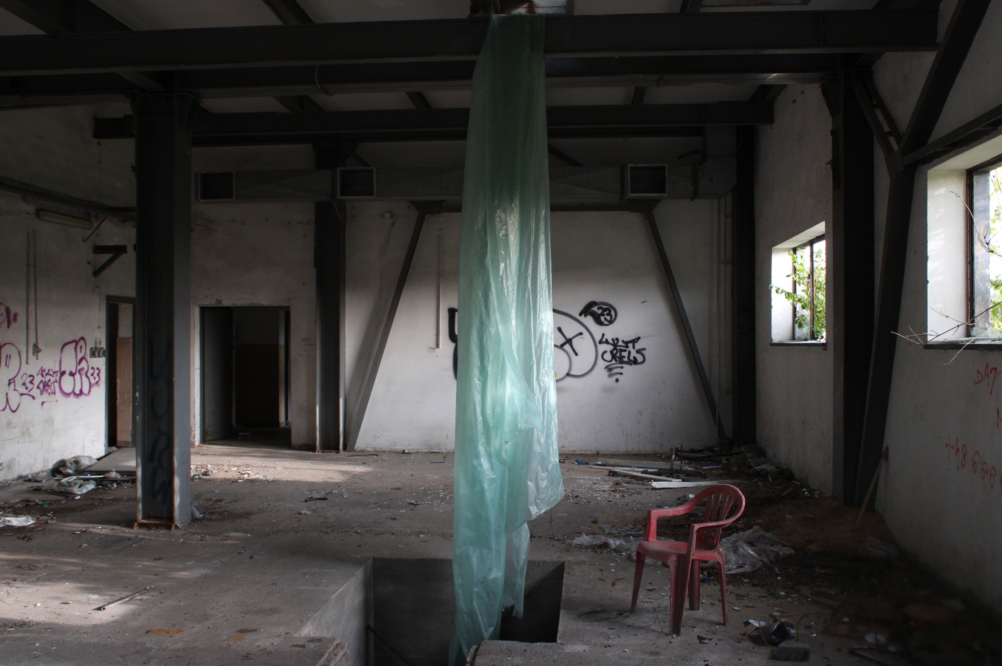 Opuszczone wnętrze hali w Wronkach z odpadami, graffiti na ścianach i plastikową folią zwisającą z sufitu, pojedyncze czerwone krzesło.