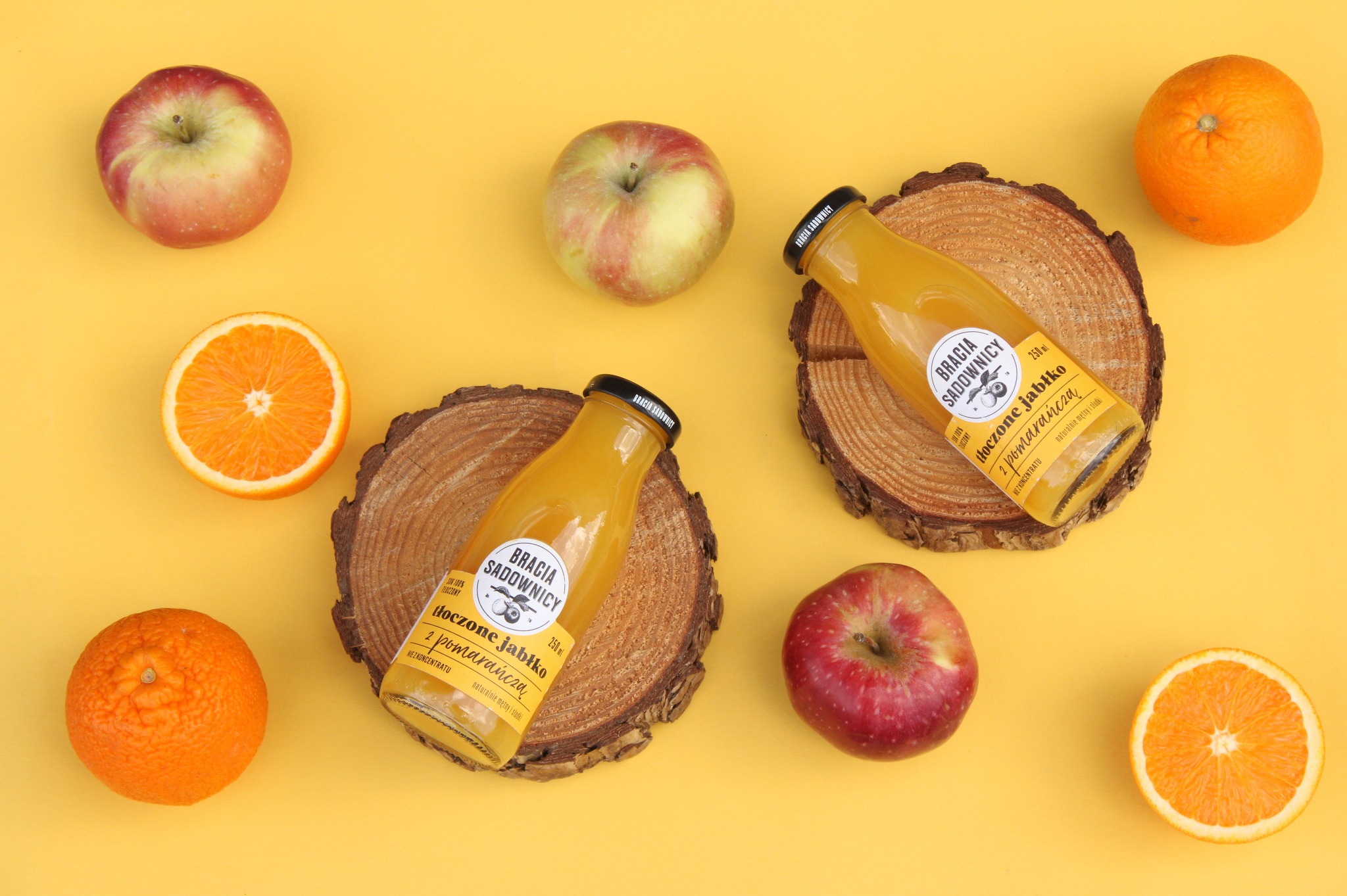 Dwie butelki soku jabłkowo-pomarańczowego Bracia Sadownicy na drewnianych podkładkach, otoczone jabłkami i pomarańczami na żółtym tle.