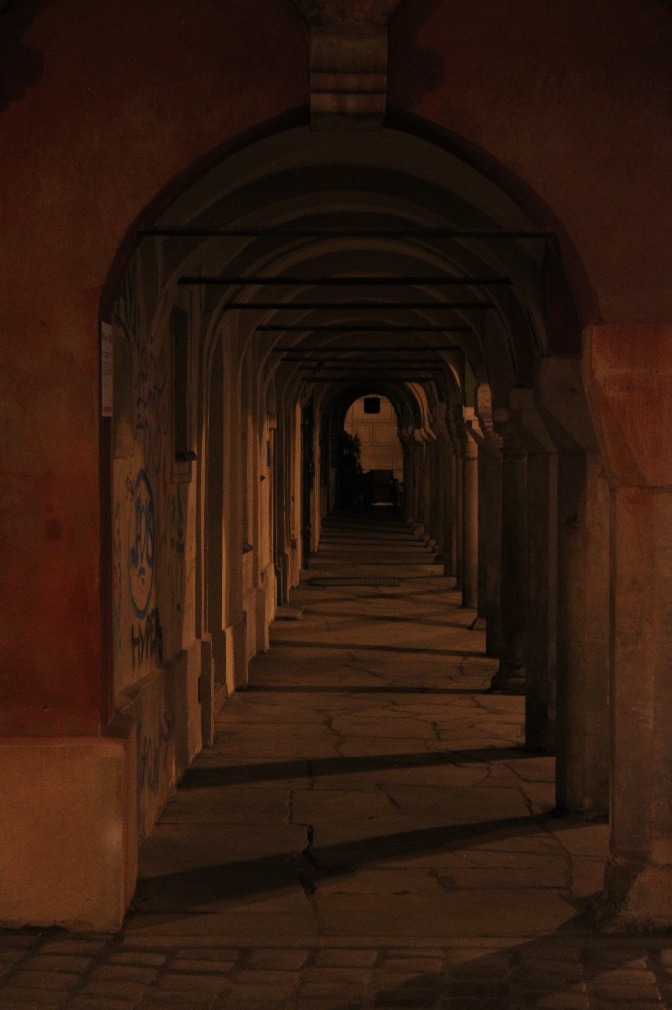 Długi, ciemny korytarz z kamiennymi kolumnami i łukami, oświetlony słabym światłem, z graffiti na ścianach po lewej stronie.