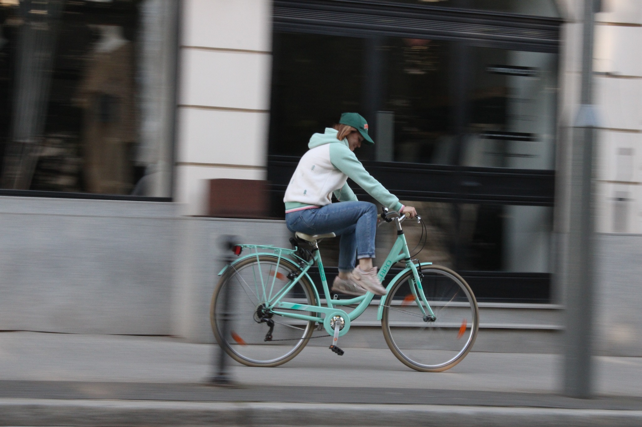 Kobieta w dżinsach i bluzie w pastelowych kolorach jedzie na turkusowym rowerze po chodniku obok budynku z dużymi oknami, w ruchu.