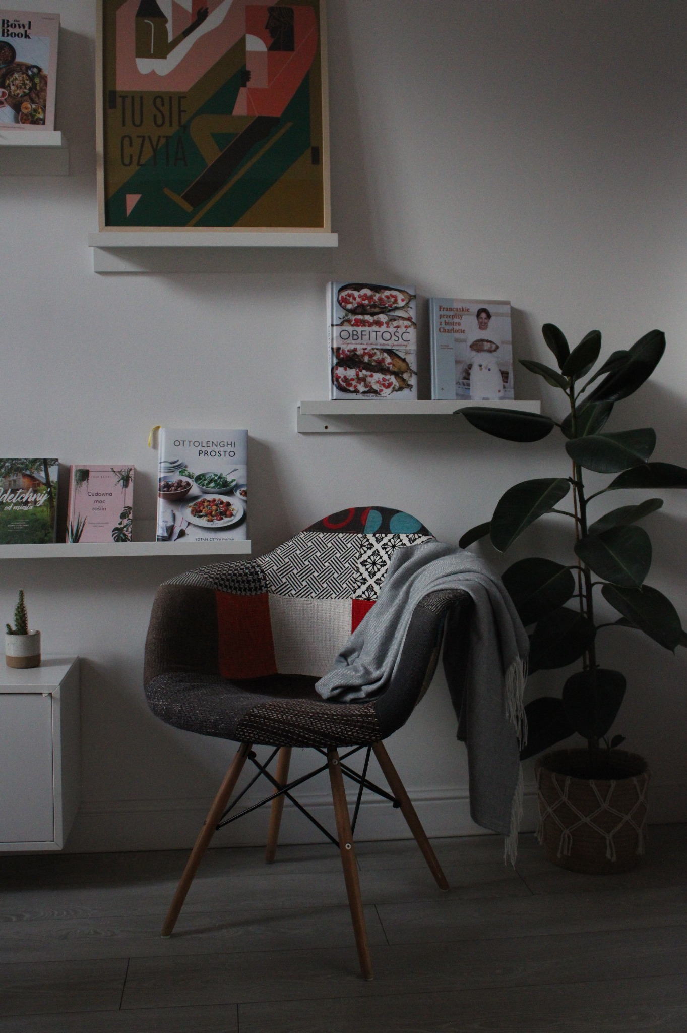 Przytulny kącik do czytania z krzesłem patchworkowym, pledem, półkami z książkami kucharskimi i plakatem 'Tu się czyta', dopełniony rośliną doniczkową w plecionym koszu.