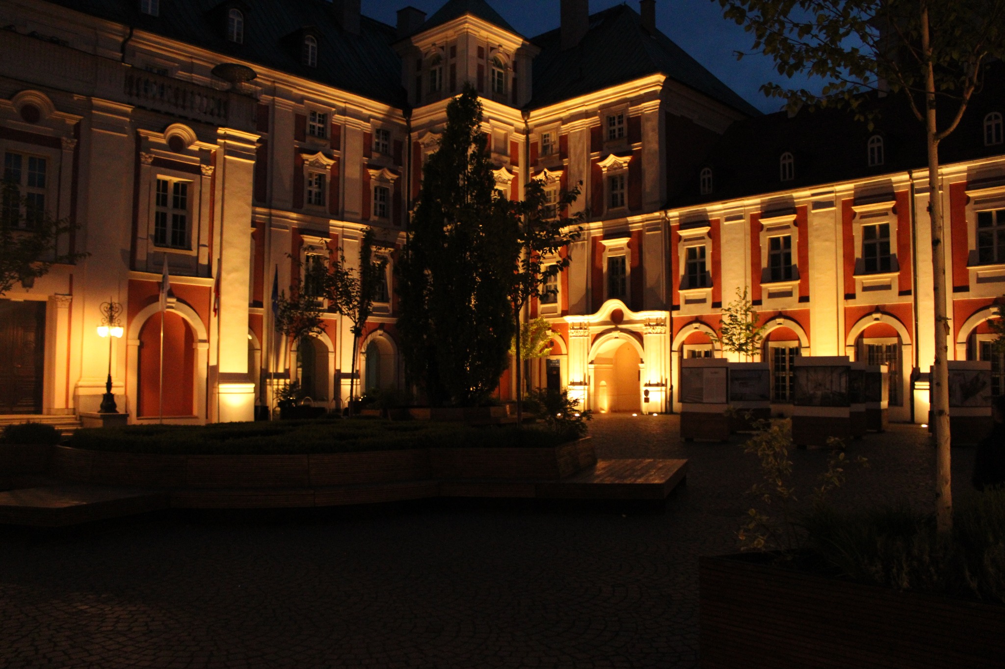 Fasada budynku w stylu barokowym, oświetlona ciepłym światłem nocą, z zielenią i brukowaną nawierzchnią na pierwszym planie.