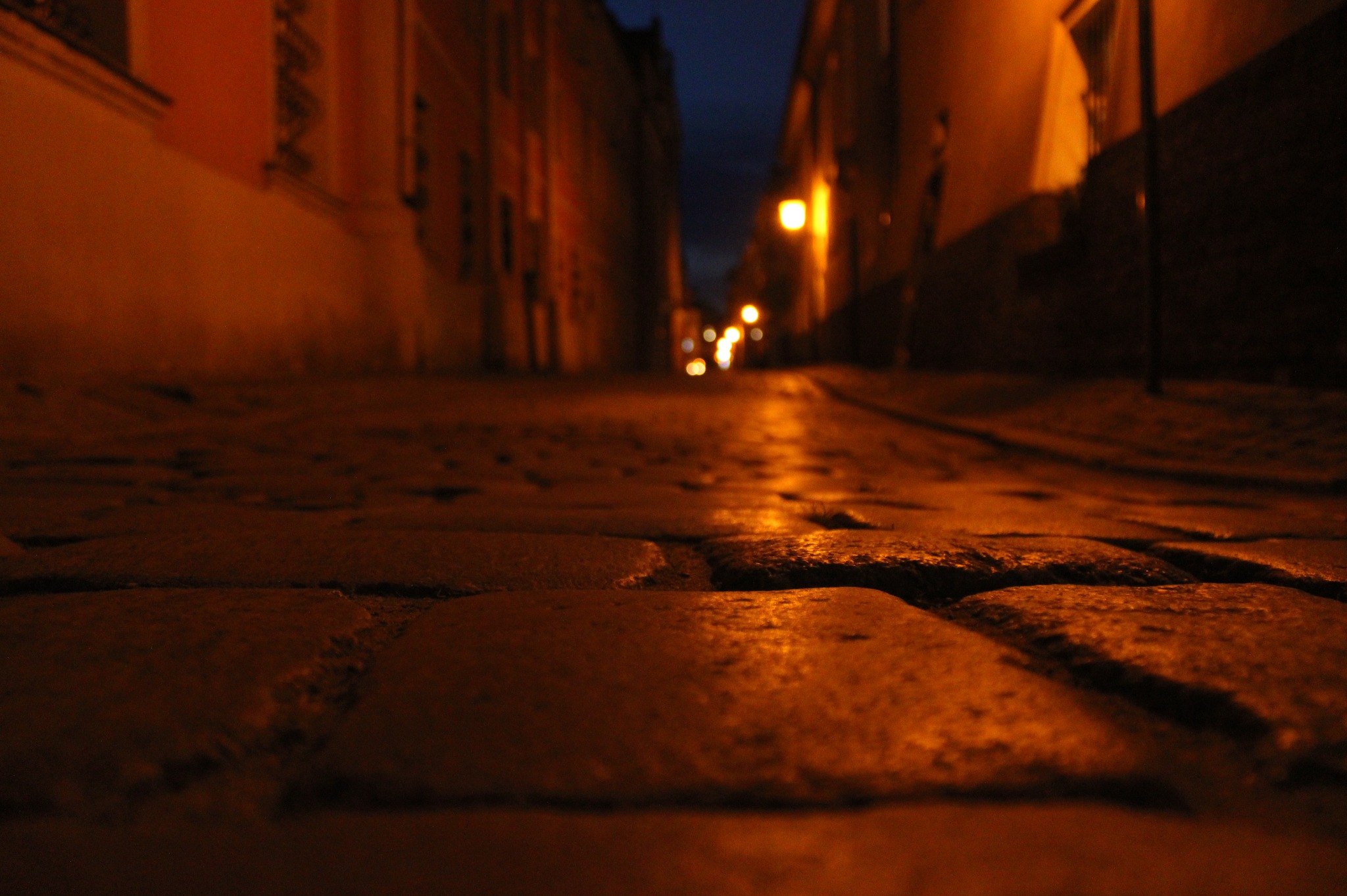 Ulica wybrukowana kostką, oświetlona ciepłym światłem latarni w nocnej scenerii miejskiej, z budynkami po obu stronach drogi.