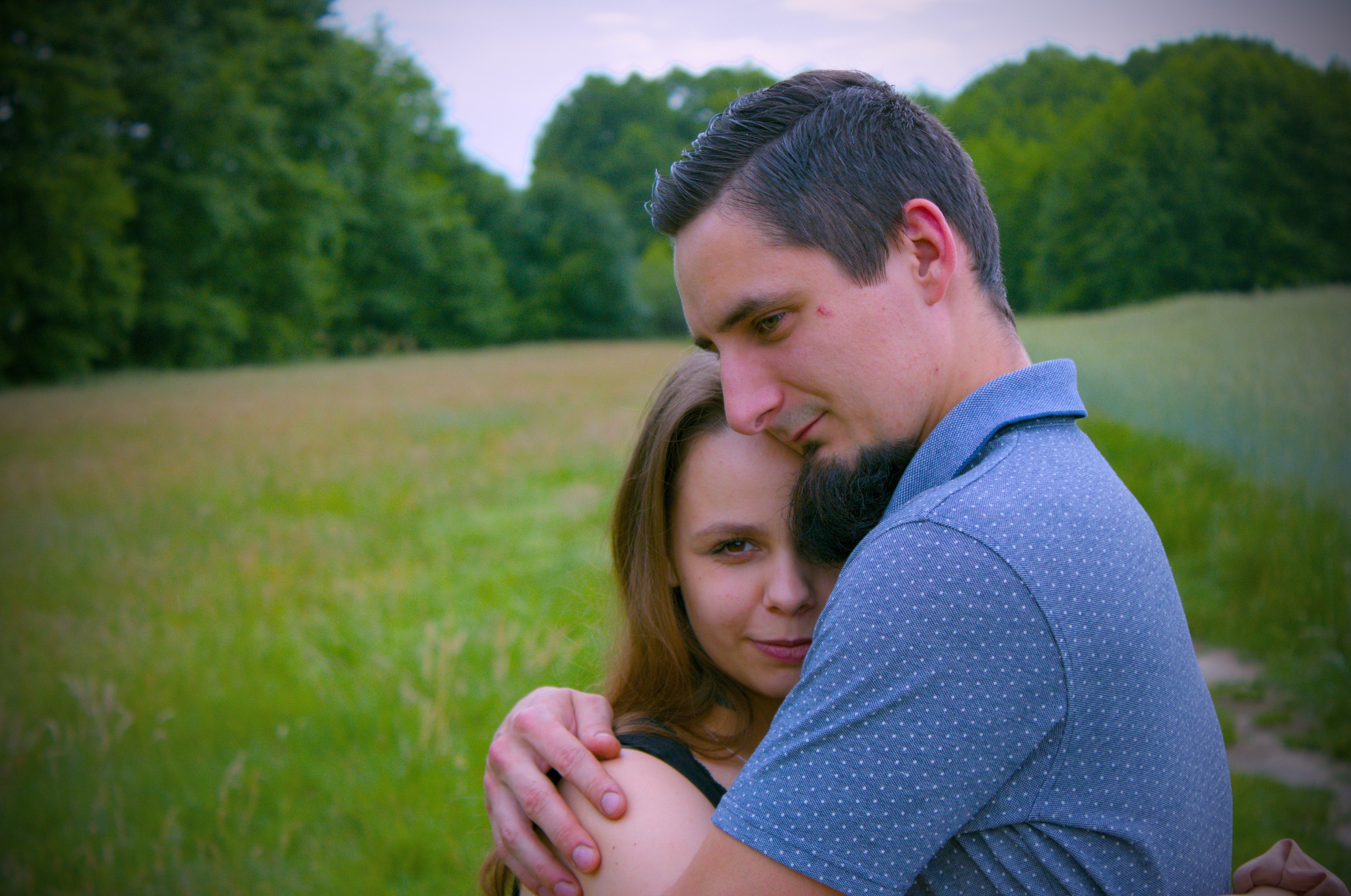 Młoda para obejmuje się na łące, mężczyzna w niebieskiej koszuli w kropki, kobieta z długimi włosami patrzy w obiektyw.