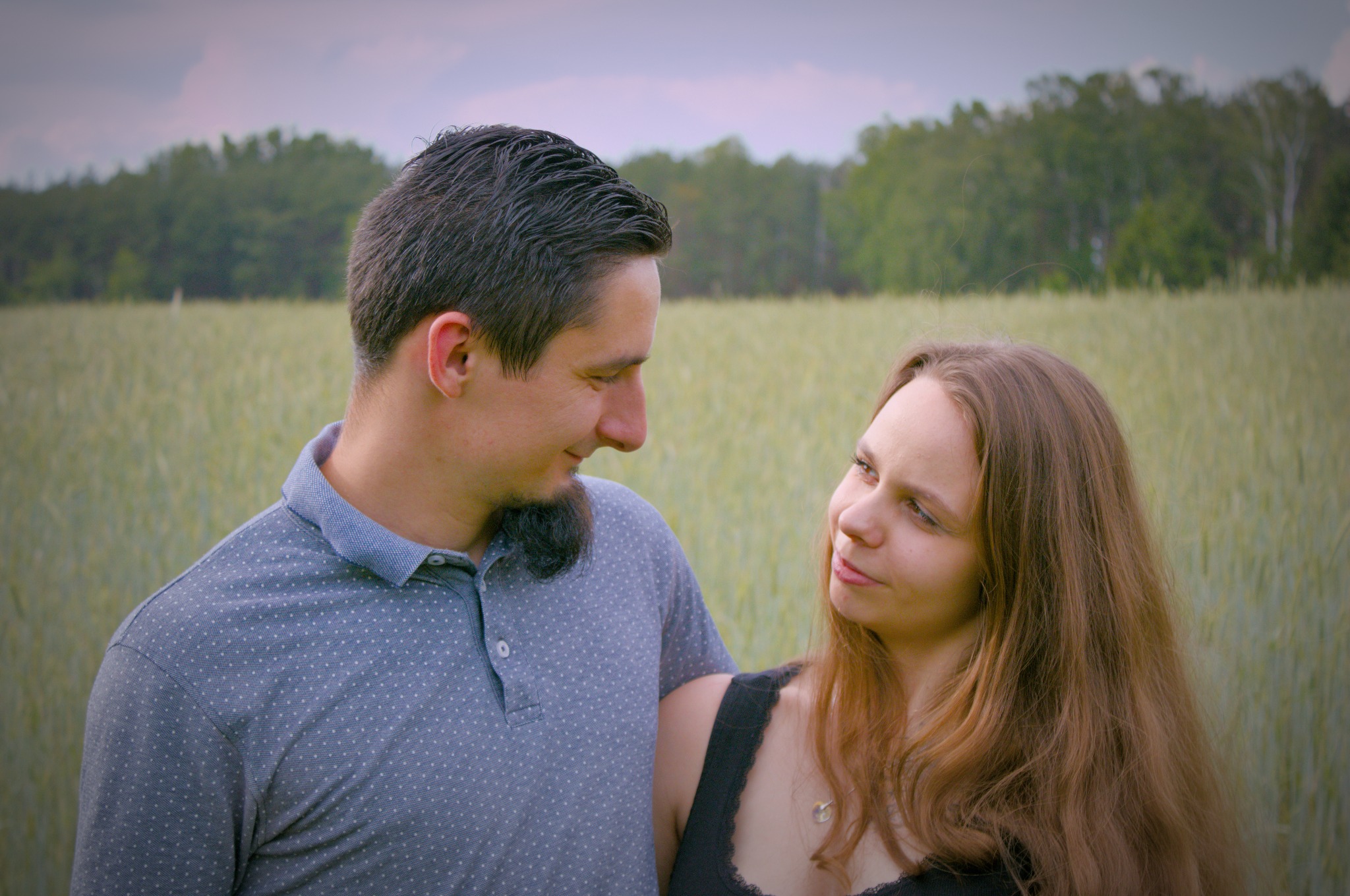 Zakochana para w polu pszenicy, mężczyzna w koszulce polo z krótkim rękawem spogląda na kobietę z długimi brązowymi włosami, w tle las.