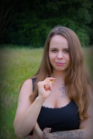 Kobieta z długimi włosami i tatuażami na ramionach, w czarnym topie, stoi na łące i trzyma w palcach mały, żółty przedmiot.