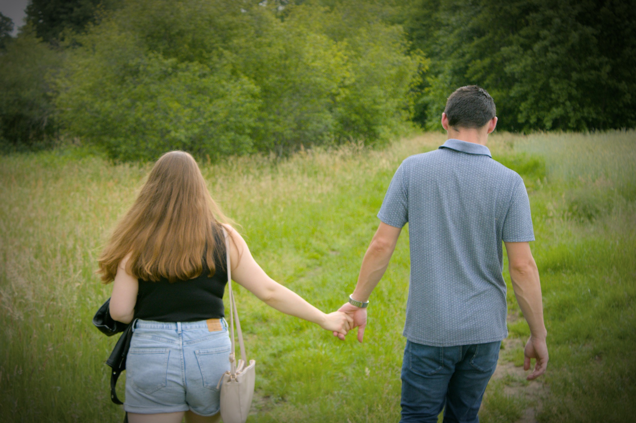 Para trzymająca się za ręce podczas spaceru po łące w okolicach Wronek, widok z tyłu, zieleń w tle.