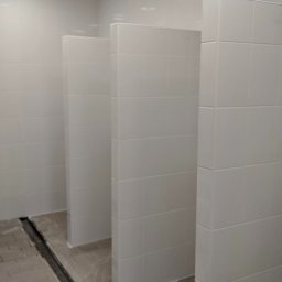 Volodymyr Tsoi - Publiczna toaleta w trakcie remontu: widoczne kabiny z białych płytek, niedokończona podłoga z odpływem liniowym, sufit z kwadratowymi lampami.