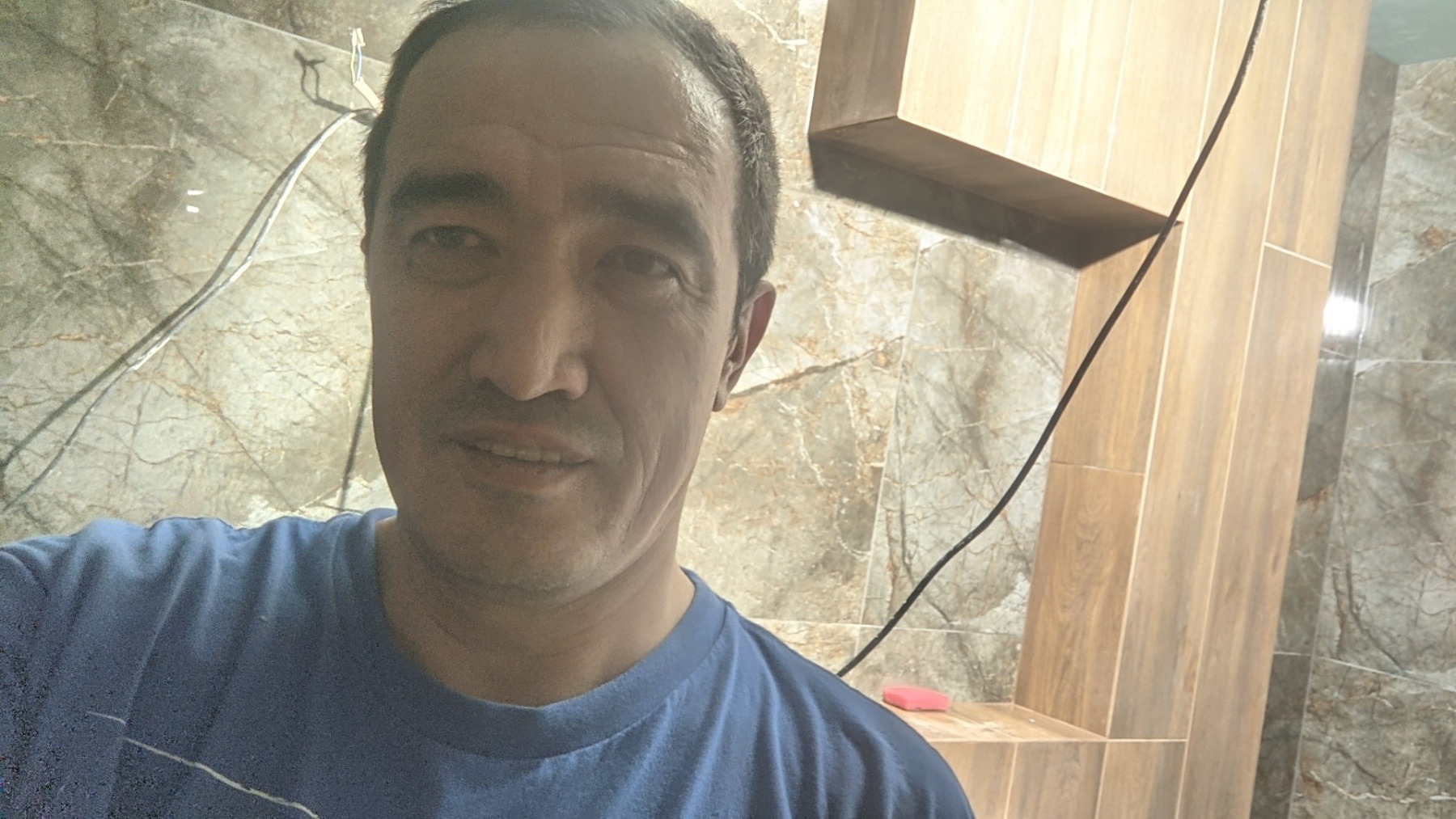 Autoportret glazurnika na tle częściowo wykończonej łazienki z marmurowymi i drewnopodobnymi płytkami, widoczne przewody elektryczne.