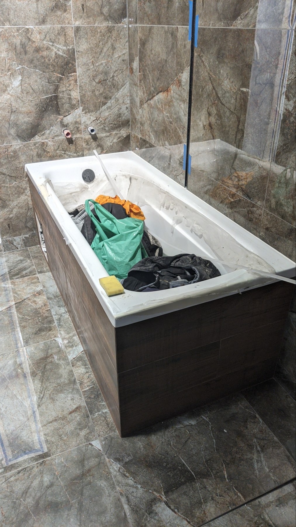 Wanna w trakcie obudowy płytkami imitującymi drewno, widoczne narzędzia i ubrania wewnątrz wanny, niedokończone fugowanie płytek na ścianach.