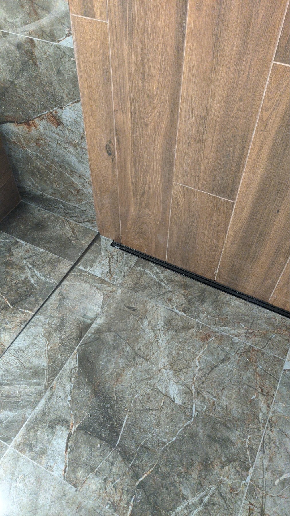 Szare marmurowe płytki podłogowe w łazience, połączone z pionowymi płytkami ściennymi imitującymi drewno, widoczny czarny profil dylatacyjny.