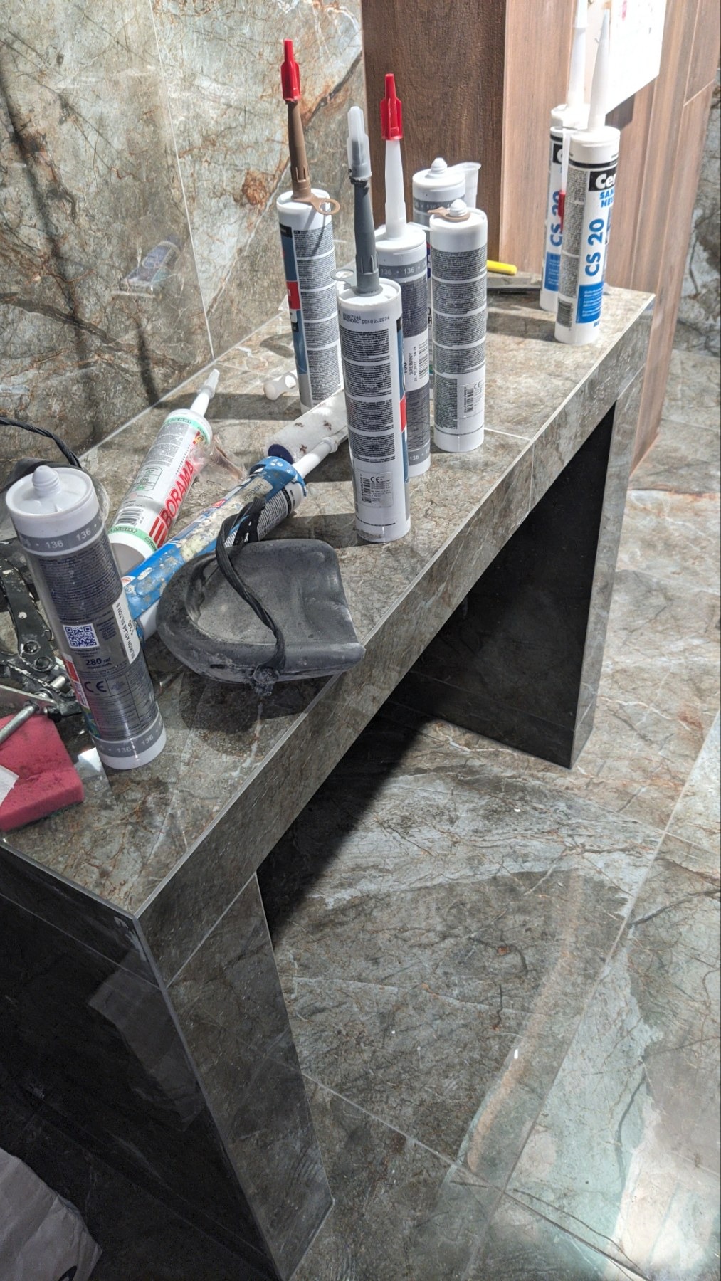 Marmurowa umywalka z narzędziami i tubami silikonu budowlanego, widoczne aplikatory z czerwonymi i szarymi końcówkami, oraz niebieska szpachelka.