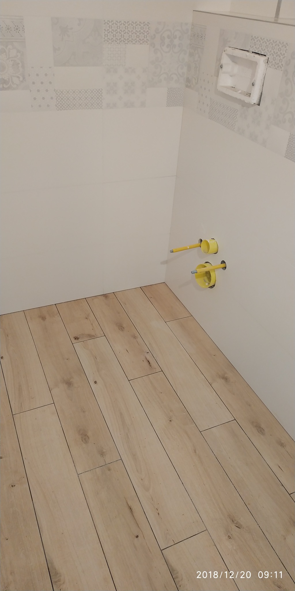 Fragment łazienki w trakcie remontu: ułożona podłoga z paneli imitujących drewno, białe płytki na ścianach z dekoracyjnym pasem u góry, widoczne żółte elementy instalacji wodnej i otwór na stelaż...