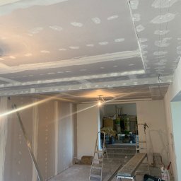 Twoje Wnętrze - Wnętrze w trakcie remontu: ściany wyłożone płytami gipsowo-kartonowymi, sufit z elementami szpachlowania, widoczne drabiny i narzędzia budowlane, przygotowania do dalszych prac wykończeniowych.