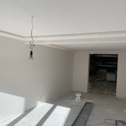 Twoje Wnętrze - Wnętrze w trakcie prac wykończeniowych: świeżo pomalowane ściany w odcieniach bieli, sufit z dekoracyjnymi elementami, wisząca żarówka na kablu, betonowa podłoga w trakcie przygotowania, widoczne...