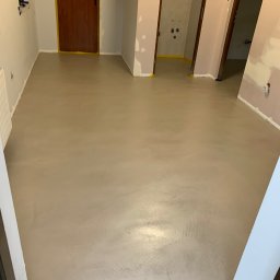 Twoje Wnętrze - Jasnoszara, gładka posadzka w trakcie prac wykończeniowych, widoczne zabezpieczone taśmą malarską ościeżnice drzwiowe i ściany w surowym stanie.