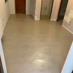 Jasnoszara, gładka posadzka w trakcie prac wykończeniowych, widoczne zabezpieczone taśmą malarską ościeżnice drzwiowe i ściany w surowym stanie.